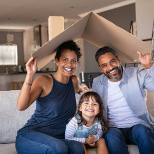 Portrait of smiling family sitting on couch holding cardboard roof and looking at camera. African and indian parents with funny daughter holding cardboard roof over heads while sitting on sofa in their new home. Happy mother and father with daughter in new house at moving day with copy space.