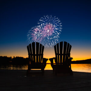 Canada day fireworks on a lake in Muskoka, Ontario Canada. On the wooden dock two Adirondack chairs are facing the sunset orange hues