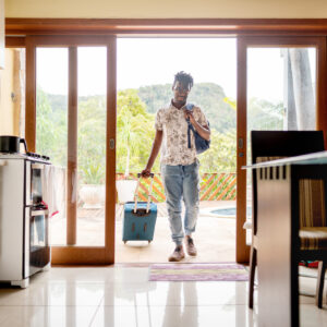 Young man walking through the patio doors of his rental accommodation pulling a wheeled suitcase